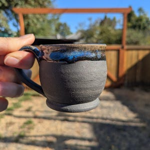 May include: A ceramic mug with a matte charcoal gray textured body and a glossy rim featuring a band of deep blue glaze transitioning into a metallic bronze tone. The mug has a rounded base and a curved handle extending from the side.