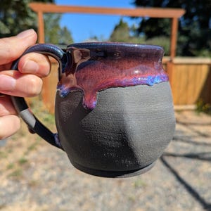 Op de afbeelding: Handgemaakte keramische mok met een getextureerde zwarte basis en een glanzend, kleurveranderend glazuur in tinten paars, rood en blauw. De mok heeft een afgeronde body en een stevig handvat.