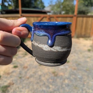 May include: A ceramic mug with a matte black base and a glossy, deep blue glaze dripping from the rim. The body features a textured, horizontal white streak across the center. The mug has a curved handle and a rounded, footed base design.