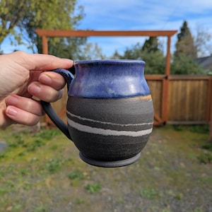 Könnte beinhalten: Handgefertigte Keramik-Tasse mit dunkelblau glasierter Oberseite und schwarzem Boden mit zwei weißen Streifen. Die Tasse hat einen schwarzen Henkel und wird vor einem unscharfen Hintergrund im Freien gehalten.