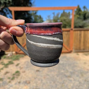 May include: A ceramic mug with a textured matte black body featuring two horizontal white wavy lines. The upper rim is glazed in a glossy deep red with a dark purple transition. The mug has a curved handle and a circular base.