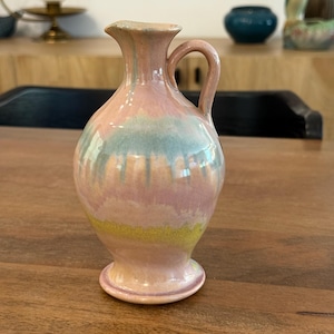 Op de afbeelding: Een kleine, pastelkleurige keramische kan met een handvat. De kan heeft een bolvormig lichaam, een smalle hals en een uitlopende rand. De kleuren zijn roze, blauw en geel, met een glanzende afwerking. De kan is waarschijnlijk voor decoratief gebruik.