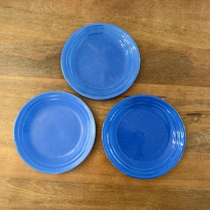 May include: Three blue ceramic plates with a slightly rippled edge. The plates are arranged in a triangular pattern on a wooden surface.