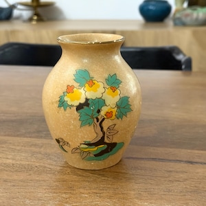 Puede incluir: Un pequeño jarrón de cerámica de color crema con un borde dorado y un diseño floral. El jarrón presenta un árbol estilizado con flores amarillas y hojas verdes.