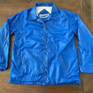 May include: A blue nylon jacket with a snap closure and two pockets. The jacket has a white lining and a small label with the text "Sun Surf" on the left side of the collar.