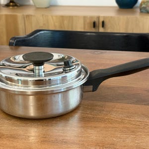 Puede incluir: Una cacerola de acero inoxidable con asa y tapa negras. La tapa tiene un pomo negro y una válvula de liberación de presión. La olla está sobre una superficie de madera. Utensilios de cocina.