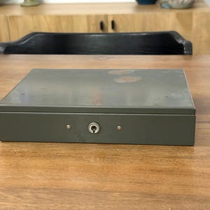 Peut inclure: Une boîte métallique rectangulaire grise avec un trou de serrure et deux petits rivets sur le devant. La boîte est fermée et posée sur une surface en bois. L'arrière-plan comprend un meuble en bois et une chaise noire.