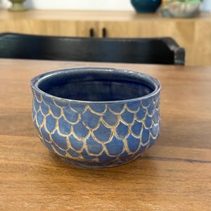 Pode incluir: Uma pequena tigela redonda de cerâmica azul com um padrão texturizado em forma de escamas em uma cor mais clara. A tigela está sobre uma superfície de madeira, mostrando sua qualidade artesanal e design único.