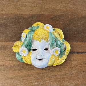 Puede incluir: Una máscara de cerámica con una cara blanca, agujeros negros para los ojos y una boca sonriente. La máscara está adornada con cabello amarillo, hojas verdes y flores blancas con centros amarillos. La máscara es probablemente decorativa.