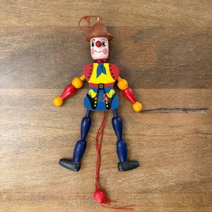 May include: A vintage wooden marionette doll with a painted clown design. The doll has a brown hat, red and yellow accents, blue pants, and black shoes. It is controlled by strings and a wooden stick.