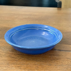 May include: A small, blue ceramic bowl with a white rim. The bowl has a slightly textured surface and is sitting on a wooden table.