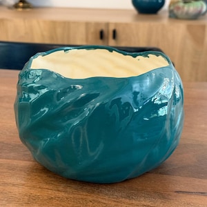 Puede incluir: Un cuenco de cerámica turquesa con un exterior texturizado y ondulado. El interior es de color crema claro. El cuenco es redondo y se apoya sobre una superficie de madera. El acabado brillante refleja la luz, resaltando la forma y el diseño únicos.