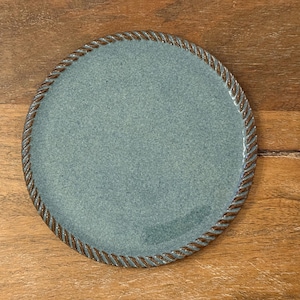 May include: A round, teal-colored ceramic plate with a textured surface and a brown rope-like border. The plate is on a wooden surface. The plate has a small handle.
