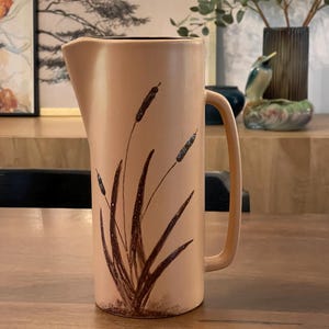 Puede incluir: Una jarra de cerámica beige con un diseño floral marrón de hierba alta y juncos. La jarra tiene un asa y una boca ancha.