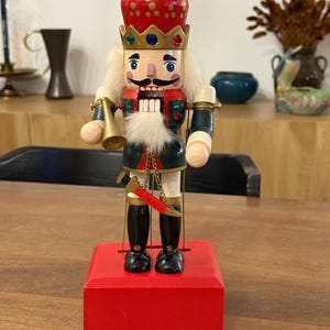 Puede incluir: Figura de cascanueces de madera con forma de rey, sobre una base roja. El cascanueces lleva una corona roja con detalles dorados, un bigote negro y una barba blanca. Sostiene una trompeta dorada y una espada.