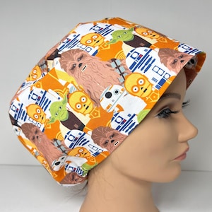 May include: A patterned surgical scrub cap featuring Star Wars characters like Chewbacca, Yoda, and R2-D2 on an orange and white background. The cap is made of fabric and is designed to cover the head.