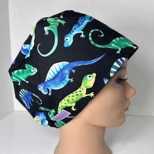 May include: A black surgical scrub cap featuring a colorful pattern of lizards in shades of green, blue, and yellow. The lizards are set against a black background. The cap is designed to fit comfortably.