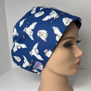 May include: A blue scrub cap featuring a repeating pattern of white owls carrying letters and brooms. The owls are depicted with open wings against a dark blue background. The cap is made of fabric and is designed to cover the head.