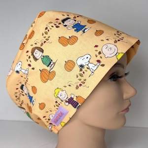 May include: A light orange scrub cap featuring a repeating pattern of Peanuts characters including Snoopy, Charlie Brown, and Lucy surrounded by pumpkins and autumn leaves. A small rectangular purple tag with white text is attached to the side.