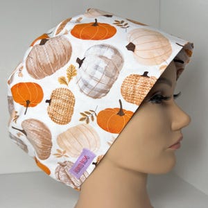 May include: A white scrub cap featuring a repeating pattern of pumpkins in shades of orange, beige, and grey, with some pumpkins displaying plaid or textured designs. A small purple rectangular tag with the text Enchanted Brims is attached.