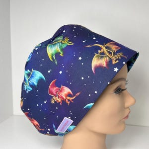 May include: A dark blue scrub cap featuring a repeating pattern of colorful dragons and white stars. The dragons are in shades of red, yellow, green, and blue. The cap is designed to fit comfortably and securely.