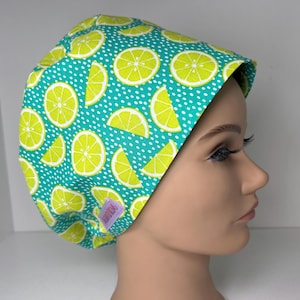 May include: A teal scrub cap with a pattern of lime slices and white polka dots. The cap is made of fabric and has a tag on the side. The cap is designed to be worn in a medical or professional setting.