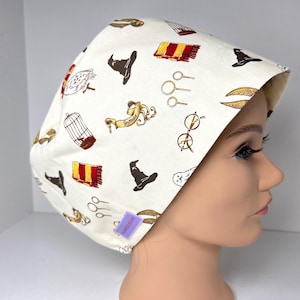 May include: A white scrub cap featuring a repeating pattern of wizarding-themed illustrations in brown, gold, and red. The design includes a wizard hat, scarf, glasses, and other iconic symbols. The cap is displayed on a mannequin head.