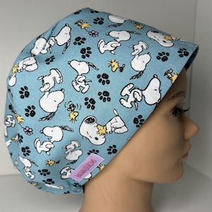 May include: A light blue scrub cap featuring a repeating pattern of white Snoopy characters, yellow Woodstock birds, small purple flowers, and black paw prints. A small rectangular purple tag with the text Enchanted Brims is attached.