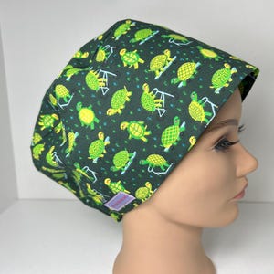 May include: A dark green scrub cap featuring a pattern of yellow and green turtles. The turtles are depicted in various poses, some with swing sets. The cap is made of a soft fabric and is designed to fit comfortably.