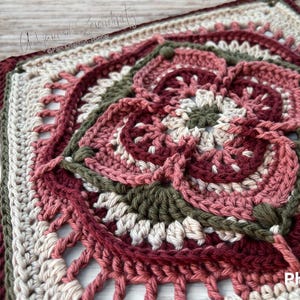 Puede incluir: Un cuadrado tejido a crochet con un diseño floral en tonos rosa, verde y crema. El cuadrado tiene un borde de hilo blanco con detalles calados.