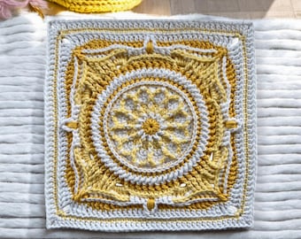 Könnte beinhalten: Ein quadratisches, gehäkeltes Kissen in Gelb- und Weißtönen. Die Mitte zeigt ein kreisförmiges Design mit einem floralen Muster. Das Kissen ist von einem weißen und gelben Muster umrandet. Das Kissen liegt auf einer weißen, strukturierten Oberfläche.