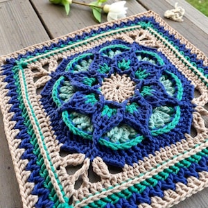 Peut inclure: Une pièce carrée au crochet faite à la main avec un motif floral. La pièce présente une fleur centrale dans les tons de bleu et de vert, entourée de bordures concentriques beiges, bleues et vertes. Le travail au crochet est détaillé et complexe.