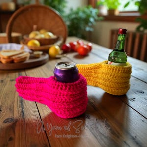 Może przedstawiać: Zbliżenie dwóch ręcznie robionych, dzierganych uchwytów na napoje. Jeden jest jasnoróżowy, trzymający fioletową puszkę, a drugi jest żółty, trzymający zieloną butelkę. Uchwyty znajdują się na drewnianym stole. Widoczny jest tekst "A Yarn of Serendipity".