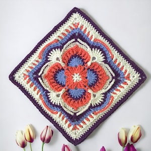 May include: A square, handmade crochet piece with a floral design. The piece features a central flower in coral, surrounded by blue, white, and purple accents. The border is a deep purple, and the piece is likely a decorative item.