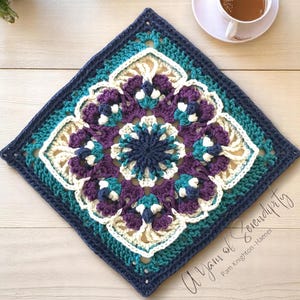 Op de afbeelding: Een vierkant, gehaakt kleedje in marine, blauwgroen, paars en crème. Het midden heeft een donker marine bloemmotief, omringd door ingewikkelde bloempatronen. Het kleedje ligt op een licht houten oppervlak. De tekst op het kleedje luidt "A Yarn of Serendipity".