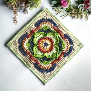 Op de afbeelding: Een vierkant, handgemaakt gehaakt stuk met een bloemenontwerp. Het stuk heeft een centrale bloem in tinten groen, rood en geel, omgeven door lagen blauw en crèmekleurig haakwerk. De randen zijn afgewerkt met een groene rand.