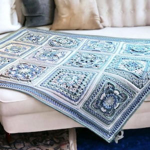 Könnte beinhalten: Eine gehäkelte Decke in Blau-, Weiß- und Grautönen mit einem quadratischen Muster mit floralen und geometrischen Designs. Die Decke ist über ein weißes Sofa drapiert und zeigt ihre komplizierten Details und Texturen. Die Gesamtästhetik ist gemütlich und handgefertigt.
