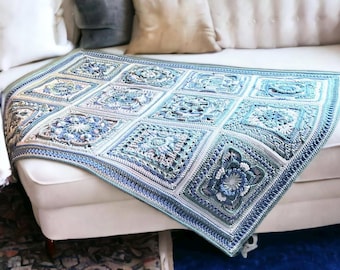 Könnte beinhalten: Eine gehäkelte Decke in Blau-, Weiß- und Grautönen mit einem quadratischen Muster mit floralen und geometrischen Designs. Die Decke ist über ein weißes Sofa drapiert und zeigt ihre komplizierten Details und Texturen. Die Gesamtästhetik ist gemütlich und handgefertigt.