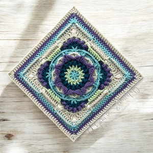 May include: A crocheted square with a colorful mandala design in shades of purple, teal, green, and white. The square is surrounded by a border of beige, blue, and purple yarn.