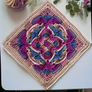 May include: A square, handmade crochet piece featuring a central floral design in shades of purple, blue, and beige. The intricate pattern is surrounded by a beige border. The yarn ball in the background suggests a craft project.