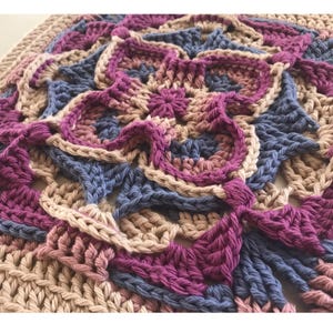 May include: Close-up of a handmade crochet blanket. The blanket features a floral design in shades of purple, blue, and beige. The crochet stitches are visible, creating a textured surface. The blanket is square-shaped and appears to be made of soft yarn.