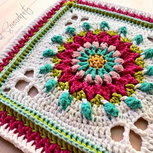 May include: A square, handmade crochet piece with a floral design. The center features a pink and yellow flower, surrounded by turquoise and pink accents. The border is white, green, and pink. The text "A Yarn of Serendipity" is in the upper left corner.