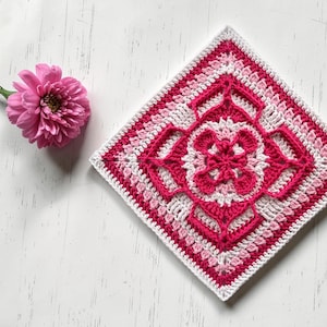May include: A square, handmade crochet piece in shades of pink and white, featuring a central floral design. The piece is surrounded by a white border. A pink flower with a green stem lies to the left of the crochet square.
