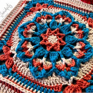 Puede incluir: Una pieza cuadrada de ganchillo hecha a mano con un diseño floral central en verde azulado, coral y crema. El intrincado patrón incluye un centro en forma de estrella y un borde detallado. El texto "a Yarn of Serendipity" es visible en la esquina superior izquierda.