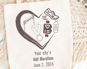 Puede incluir: Una bolsa de lona blanca con un diseño de corazón marrón que presenta iconos relacionados con la carrera, como una línea de meta, una medalla, una zapatilla para correr, una botella de agua y un monitor de frecuencia cardíaca. El texto "Your city's Half Marathon June 2, 2024" está impreso debajo del corazón.