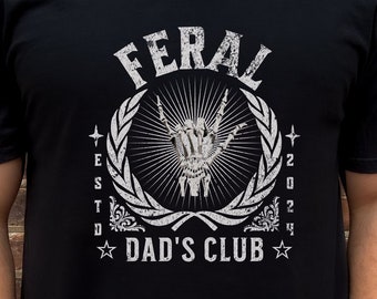 May include: Black t-shirt with a white graphic design. The graphic features a hand making the 'rock on' sign, a laurel wreath, and the text "FERAL ESTD 2024 DAD'S CLUB".