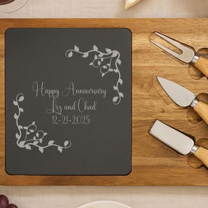 May include: A wooden cheese board with a dark gray slate insert, engraved with "Happy Anniversary Liz and Chad 12-21-2025" and floral accents. Includes three cheese knives and a fork. The board is surrounded by cheese, bread, and wine.