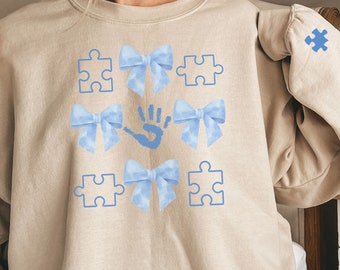 May include: A light beige sweatshirt with a blue puzzle piece design. The design features blue puzzle pieces, blue bows, and a blue handprint. The sweatshirt has a blue puzzle piece patch on the sleeve.