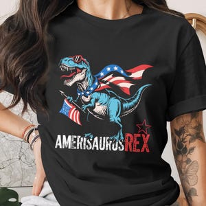 May include: Black t-shirt featuring a blue dinosaur wearing sunglasses and a patriotic cape, holding an American flag. The text "AMERISAURUS REX" is printed below the dinosaur in white and red.
