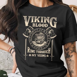 Puede incluir: Camiseta negra con temática vikinga. La camiseta presenta el texto "VIKING BLOOD RUNS THROUGH MY VEINS" en una fuente estilizada, junto con un barco vikingo, hachas y espadas. El diseño es de color crema.